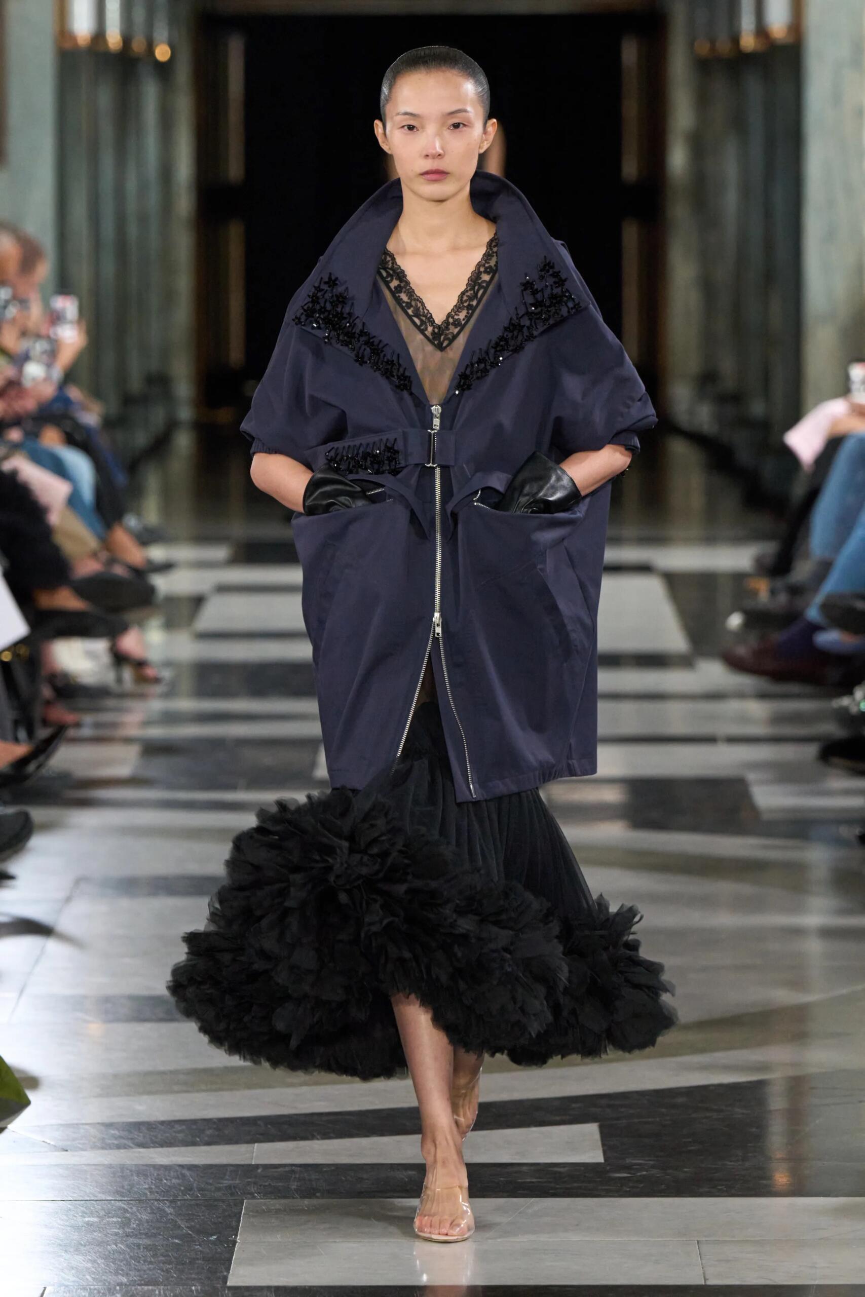 Simone Rocha printemps-été 2027, look 1 : Anorak bleu nuit, transparence et volume noir fleuri en bas de silhouette.