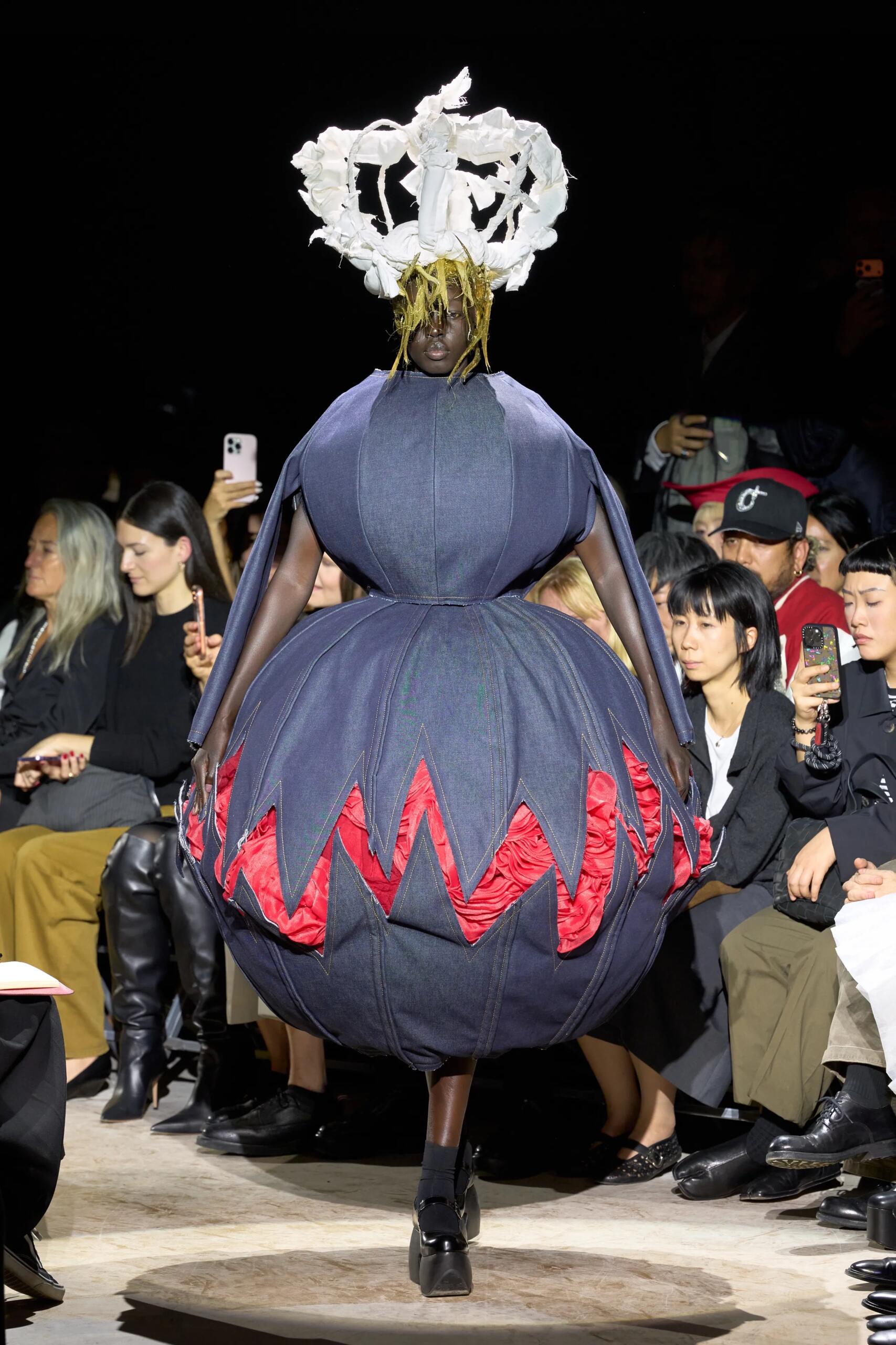 Comme des Garçons printemps-été 2027, look 1 : Volumes sphériques en denim, découpes rouges et coiffe blanche sculpturale.