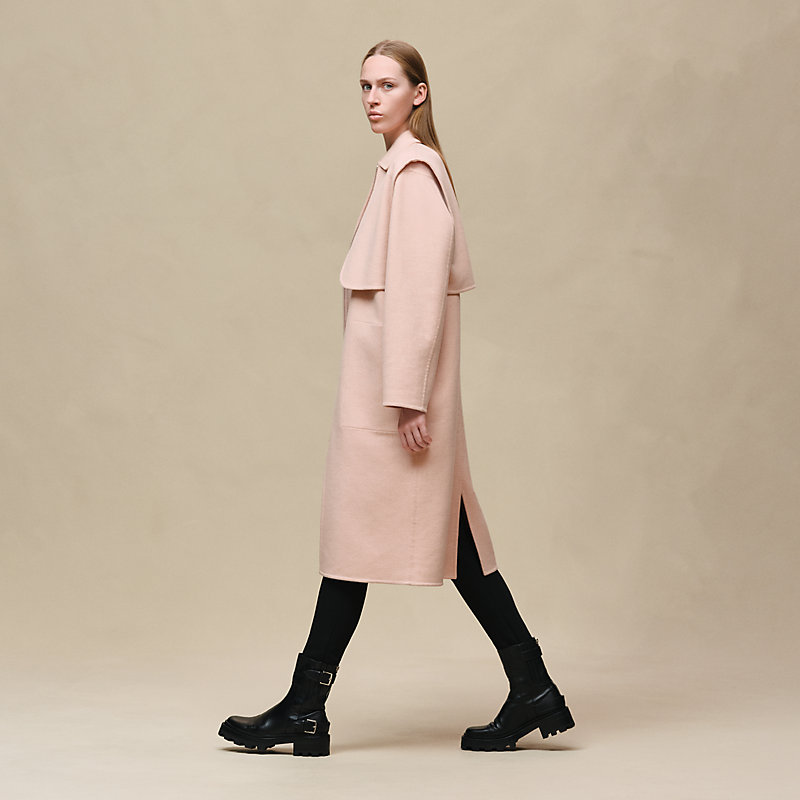 Side view of the Hermès Long storm flap coat in Rose Palomino, worn with black trousers and boots.